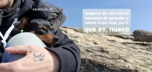 Autorretrato con Chispa escondida tras mi mano con tatto de OM. Al lado una roca y cielo azul, con caja de texto "empezar de cero con la intención de aprender, valorar lo que llega, por lo que es, nuevo" por AsunAdá