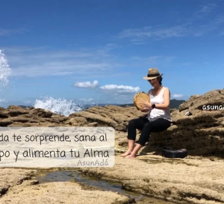 Asun Adá sentada en una roca junto al mar, con sombrero y tocando la pandereta con caja de texto: la Vida te sorprende, sana al Cuerpo y alimenta tu Alma, por AsunAdá