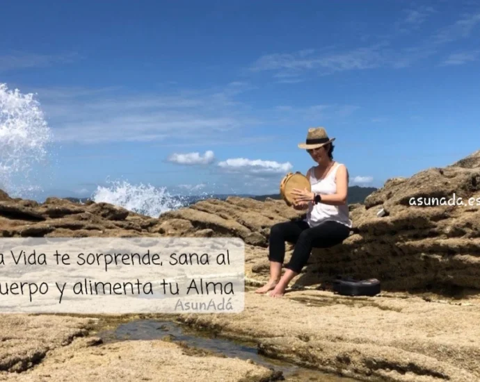 Asun Adá sentada en una roca junto al mar, con sombrero y tocando la pandereta con caja de texto: la Vida te sorprende, sana al Cuerpo y alimenta tu Alma, por AsunAdá