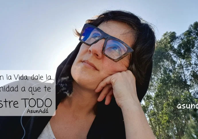 Autorretrato Asun Adá, mirando al cielo con gafas y capucha negra, detrás unos eucaliptos altos con caja de texto: confía en la Vida, dale la oportunidad a que te muestre TODO, por AsunAdá